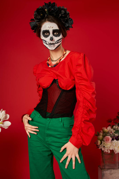 woman in sugar skull makeup and black wreath near flowers on red, dia de los muertos tradition - Photo, Image
