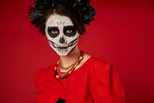 portrait of woman in sugar skull makeup and black wreath looking at camera on red, Day of Dead - Photo, Image
