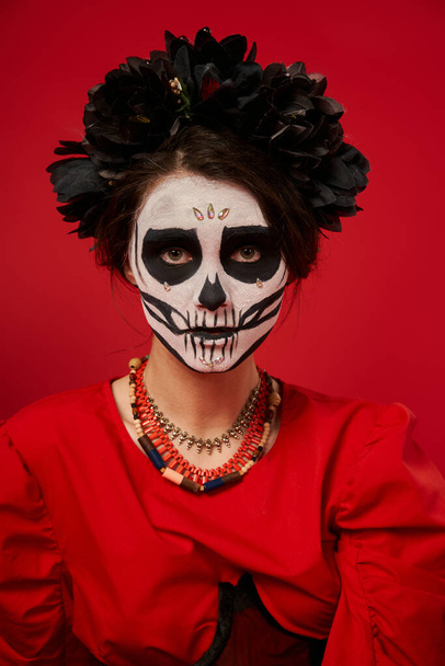 mujer en día de los muertos maquillaje y corona negra con cuentas de colores mirando a la cámara en rojo - Foto, imagen