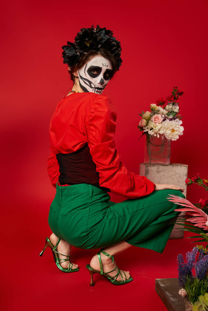 woman in dia de los muertos makeup looking at camera near traditional altar with flowers on red - Photo, Image