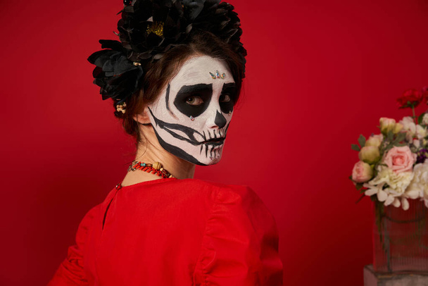 portrait of young woman in spooky catrina makeup and black wreath looking at camera on red - Foto, afbeelding