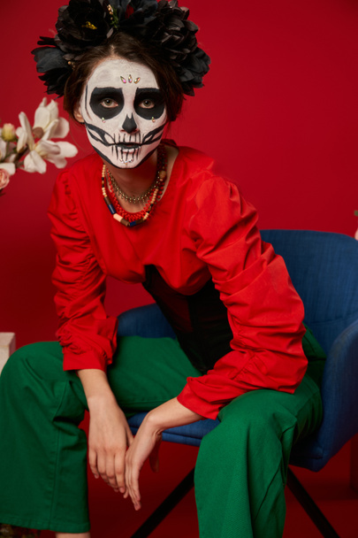 woman in dia de los muertos makeup sitting in armchair near flowers and looking at camera on red - Fotografie, Obrázek