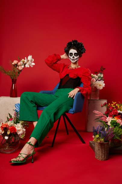 жінка в макіяжі Dia de los muerts, сидить у кріслі біля традиційного вівтаря з квітами на червоному - Фото, зображення