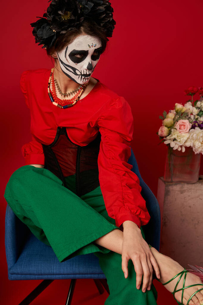 Frau in Catrina Make-up sitzt im Sessel neben Blumen auf rot, dia de los muertos tradition - Foto, Bild