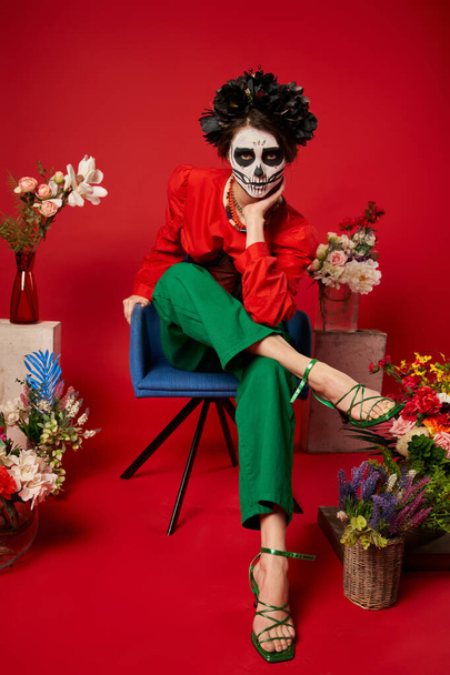 femme dans le maquillage du crâne en regardant la caméra près de dia de los muertos ofrenda avec des fleurs sur rouge - Photo, image