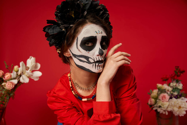 Frau in gespenstischem Totenkopf-Make-up und schwarzem Kranz blickt in die Kamera neben Blumen auf Rot, Porträt - Foto, Bild