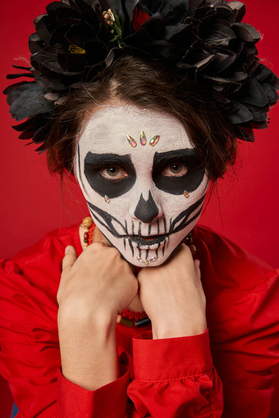 mujer en espeluznante maquillaje de cráneo y corona negra mirando a la cámara en rojo, concepto de día de los muertos - Foto, imagen