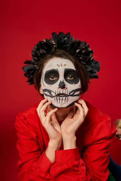 spooky woman in skull makeup and black wreath holding hands near face on red, dia de los muertos - Photo, Image