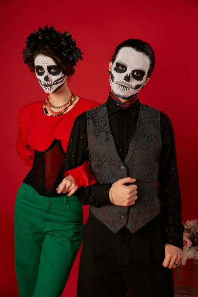 νεαρό ζευγάρι με τρομακτικό μακιγιάζ σκελετού και κομψά ρούχα ποζάρουν στο κόκκινο, dia de los muertos festival - Φωτογραφία, εικόνα