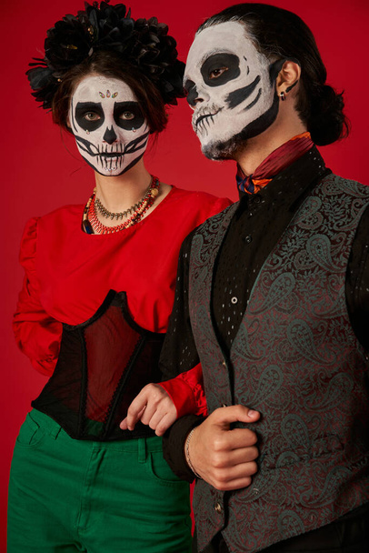 woman in skull makeup and black wreath looking at camera near man on red, dia de los muertos couple - Foto, imagen