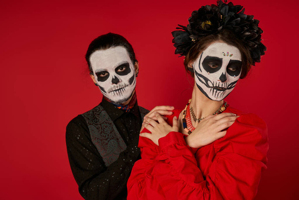 donna in trucco cranio e ghirlanda nera posa con le braccia incrociate vicino uomo spettrale sul rosso, Giorno dei morti - Foto, immagini