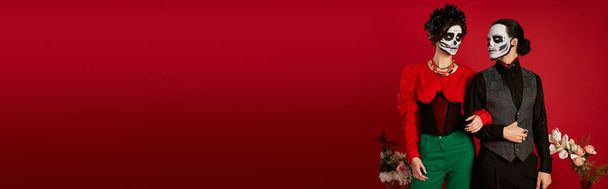 casal elegante na maquiagem catrina posando perto de flores no vermelho, dia de los muertos festival, banner - Foto, Imagem