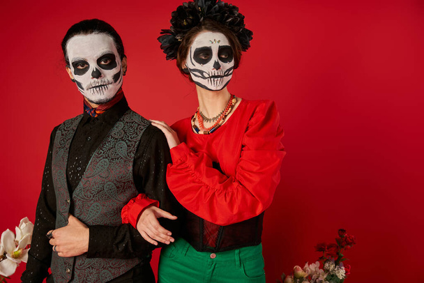 stylish couple in traditional dia de los muertos makeup looking away near flowers on red backdrop - Φωτογραφία, εικόνα