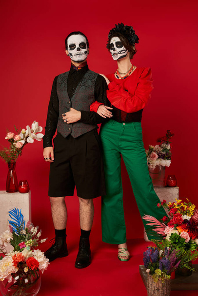 full length of couple in skull makeup near traditional Day of Dead ofrenda with flowers on red - Foto, immagini