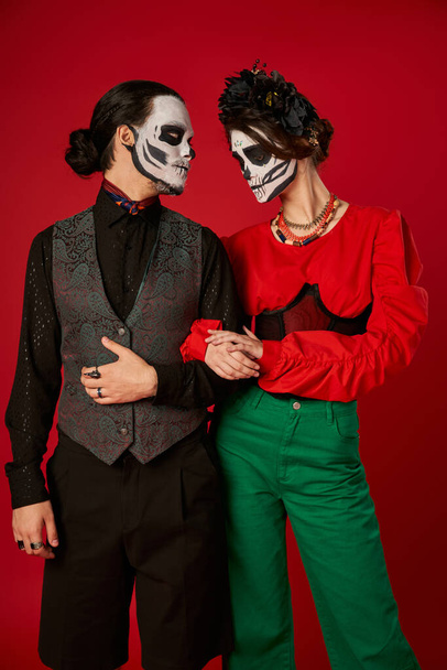 couple in trendy attire and skeleton makeup on red, dia de los muertos celebration - Foto, afbeelding