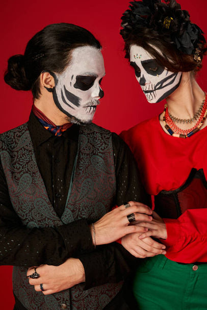 trendy couple in sugar skull makeup touching hands of each other on red, dia de los muertos festival - Fotografie, Obrázek