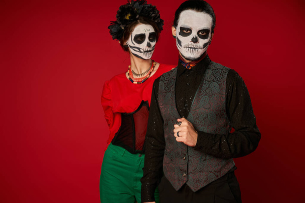 άντρας με μακιγιάζ Κατρίνα κοιτάζοντας την κάμερα κοντά σε γυναίκα με μαύρο στεφάνι, dia de los muertos ζευγάρι στο κόκκινο - Φωτογραφία, εικόνα