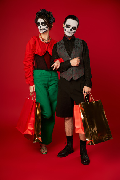 full length of trendy couple in sugar skull makeup holding shopping bags on red, dia de los muertos - Foto, immagini