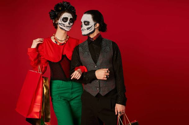 spooky couple in dia de los muertos makeup standing with shopping bags on red, festive seasonal sale - Фото, изображение