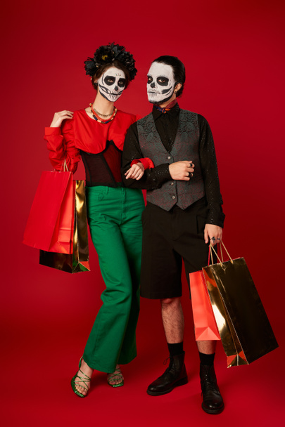 full length of couple in dia de los muertos makeup standing with shopping bags on red, festive sale - Fotoğraf, Görsel