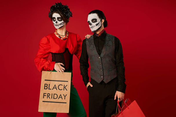 stylish couple in dia de los muertos skull makeup standing with shopping bags on red, black friday - Photo, Image