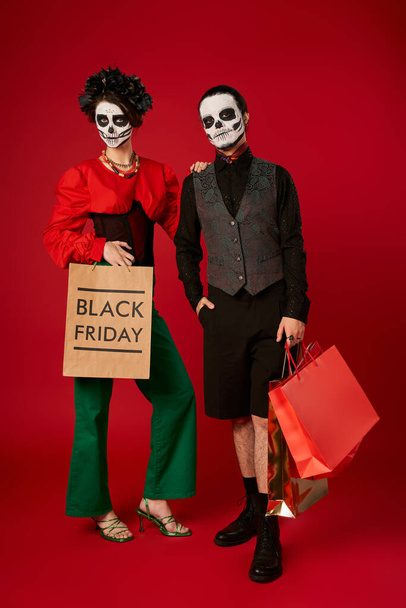full length of fashionable couple in sugar skull makeup holding shopping bags on red, black friday - Photo, Image
