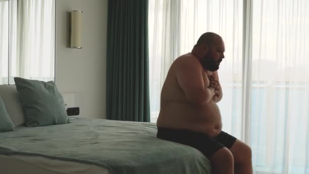 Overweight man feeling bad having seizure, difficulty breathing sitting in bed in hotel room. Exhausted guy falling in bed. Heart attack, stroke, obesity, excess weight people health problems concept. - Footage, Video