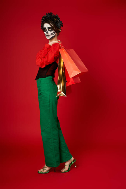 full length of woman in Day of Dead makeup and festive attire posing with shopping bags on red - Photo, Image