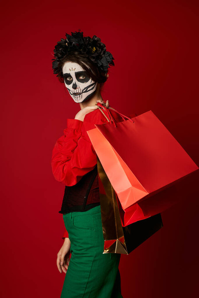 stylische Frau in schwarzem Kranz und Catrina-Make-up mit Einkaufstaschen und Blick in die Kamera auf rot - Foto, Bild