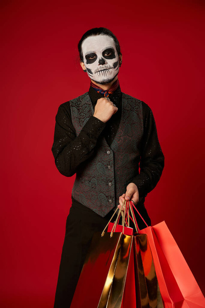 man in scary sugar skull makeup and festive attire with shopping bags on red, seasonal sale - Photo, Image