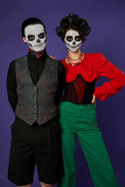 стильная пара в праздничной одежде и dia de los muertos макияж глядя на камеру на синем фоне - Фото, изображение