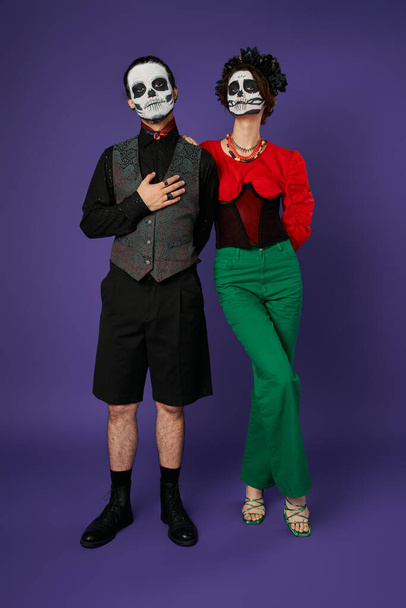 Los Muertos çifti, şeker kafa makyajlı bir adam, göğsünde bir el, mavi üzerinde ürkütücü bir kadının yanında. - Fotoğraf, Görsel