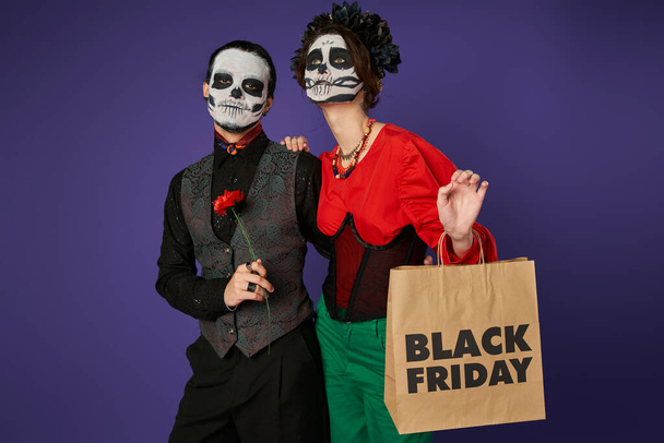 femme effrayante avec sac à provisions vendredi noir regardant loin près de l'homme dans le maquillage du crâne de sucre sur bleu - Photo, image