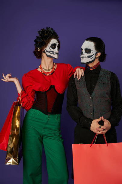 dia de los muertos couple dans le maquillage catrina tenant des sacs à provisions et se regardant sur bleu - Photo, image