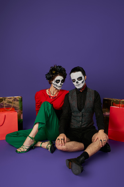 día de los muertos pareja en espeluznante esqueleto maquillaje sentado cerca de bolsas de compras en azul, venta de temporada - Foto, imagen