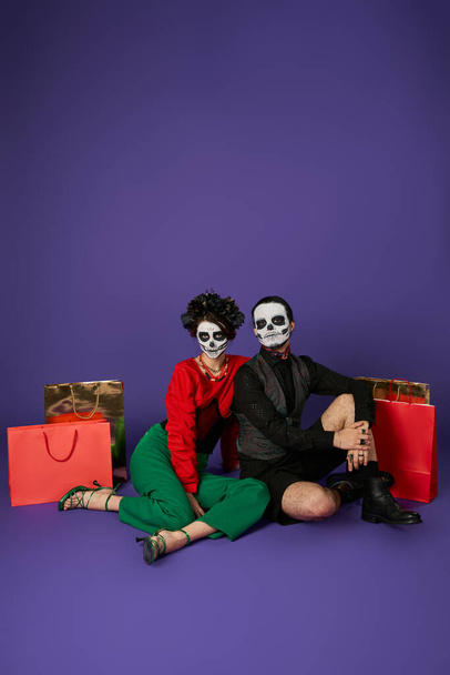 dia de los muertos couple dans effrayante squelette maquillage assis près de sacs à provisions sur bleu, pleine longueur - Photo, image