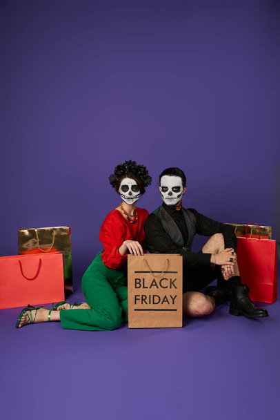 coppia alla moda in dia de los muertos cranio trucco seduto vicino a borse della spesa su blu, venerdì nero - Foto, immagini