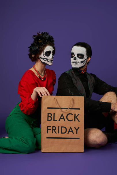 giovane coppia in tradizionale dia de los muertos trucco seduto vicino nero venerdì shopping bag su blu - Foto, immagini