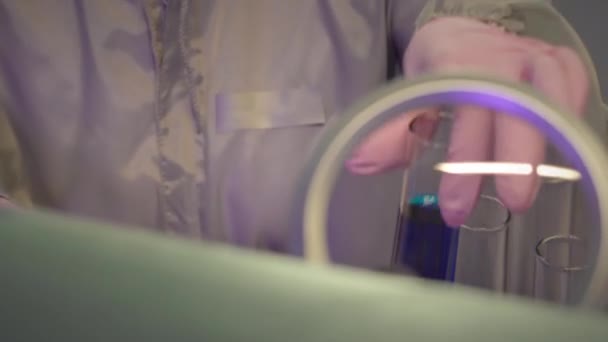 Jonge blanke vrouwelijke chemicus in beschermende kleding kijkend naar glazen reageerbuis met onherkenbare blauwe vloeistof - Video