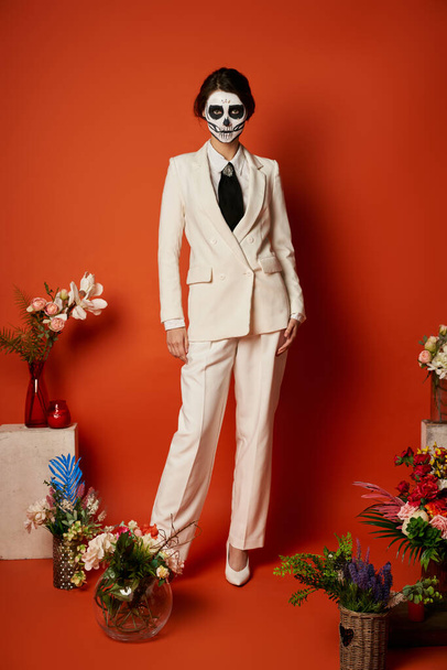 woman in skull makeup and white suit near traditional dia de los muertos ofrenda with flowers on red - Photo, Image