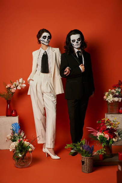couple in catrina makeup and suits posing near dia de los muertos ofrenda with flowers on red - Photo, Image