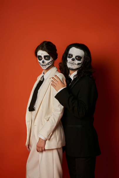κομψό ζευγάρι στο dia de los muertos μακιγιάζ και κομψά εορταστικά κοστούμια κοιτάζοντας την κάμερα στο κόκκινο - Φωτογραφία, εικόνα