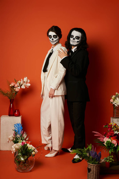 couple in skeleton makeup and suits near festive dia de los muertos ofrenda with flowers on red - Photo, Image