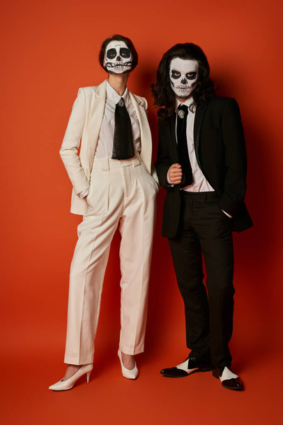 volle Länge des stilvollen Paares in Totenkopf-Make-up und eleganten Anzügen posiert mit Händen in Taschen auf rot - Foto, Bild