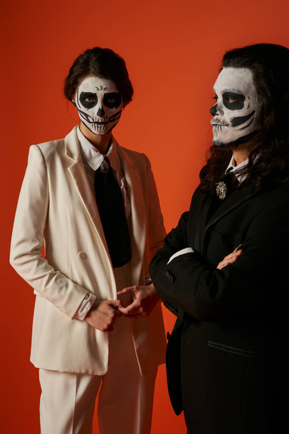 pareja en día de los muertos maquillaje, mujer en traje blanco cerca de hombre aterrador con los brazos cruzados en rojo - Foto, Imagen