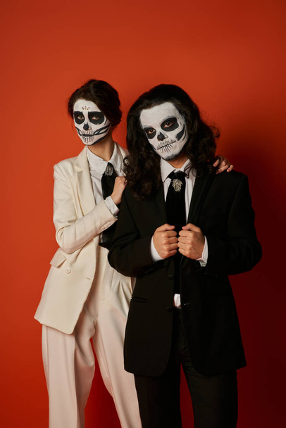 stylish couple in dia de los muertos sugar skull makeup and suits looking at camera on red backdrop - Photo, Image