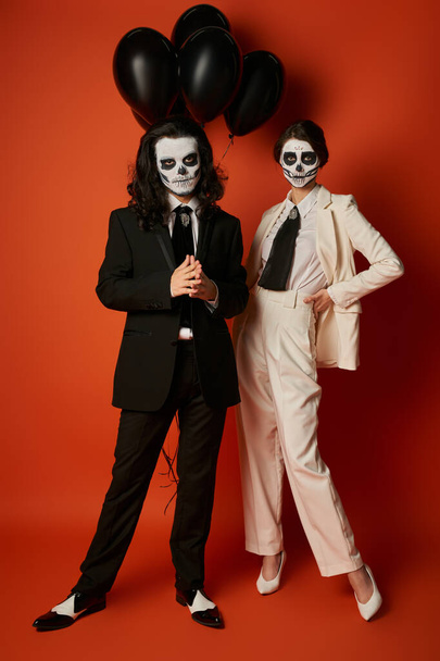 Paar in stylischen Anzügen und Catrina Calavera Make-up posiert in der Nähe schwarzer Luftballons auf Rot - Foto, Bild