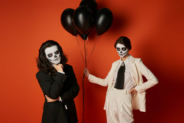 woman in skull makeup and white suit with black balloons near spooky man on red, dia de los muertos - Photo, Image