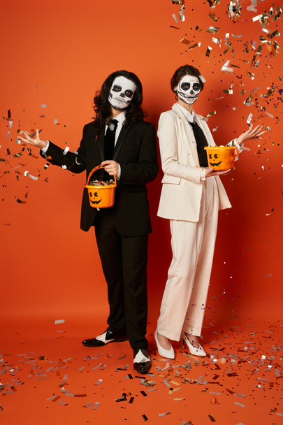 couple in skull makeup throwing shiny confetti from candy buckets on red, dia de los muertos party - Photo, Image