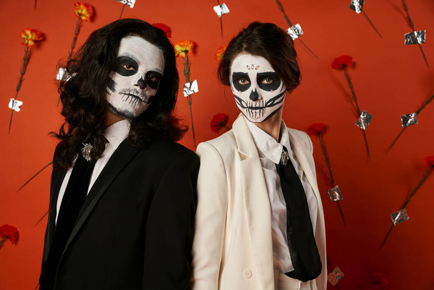 trendy couple in dia de los muertos skull makeup looking at camera on red backdrop with flowers - Fotografie, Obrázek